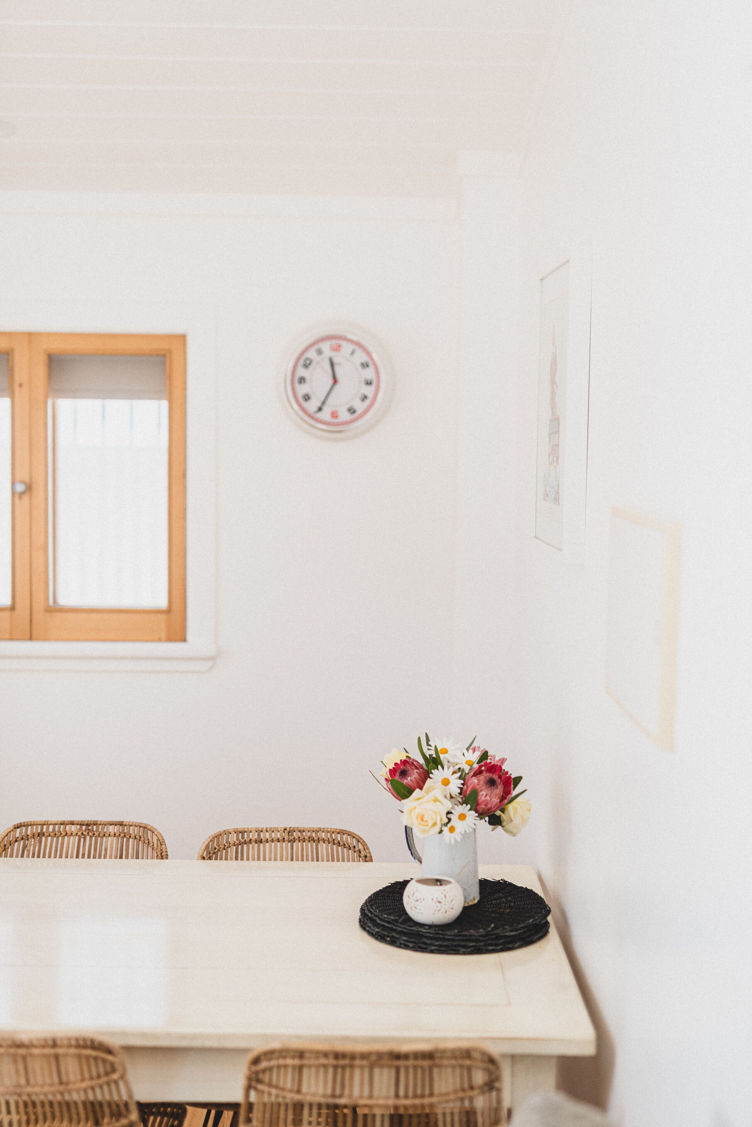 Un coin repas avec une table blanche, des chaises en osier, une horloge et un vase de fleurs.