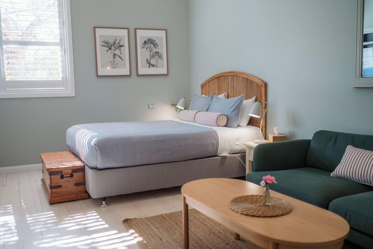 A bedroom with a bed, a green sofa, a wooden chest, and two framed botanical prints on the wall.