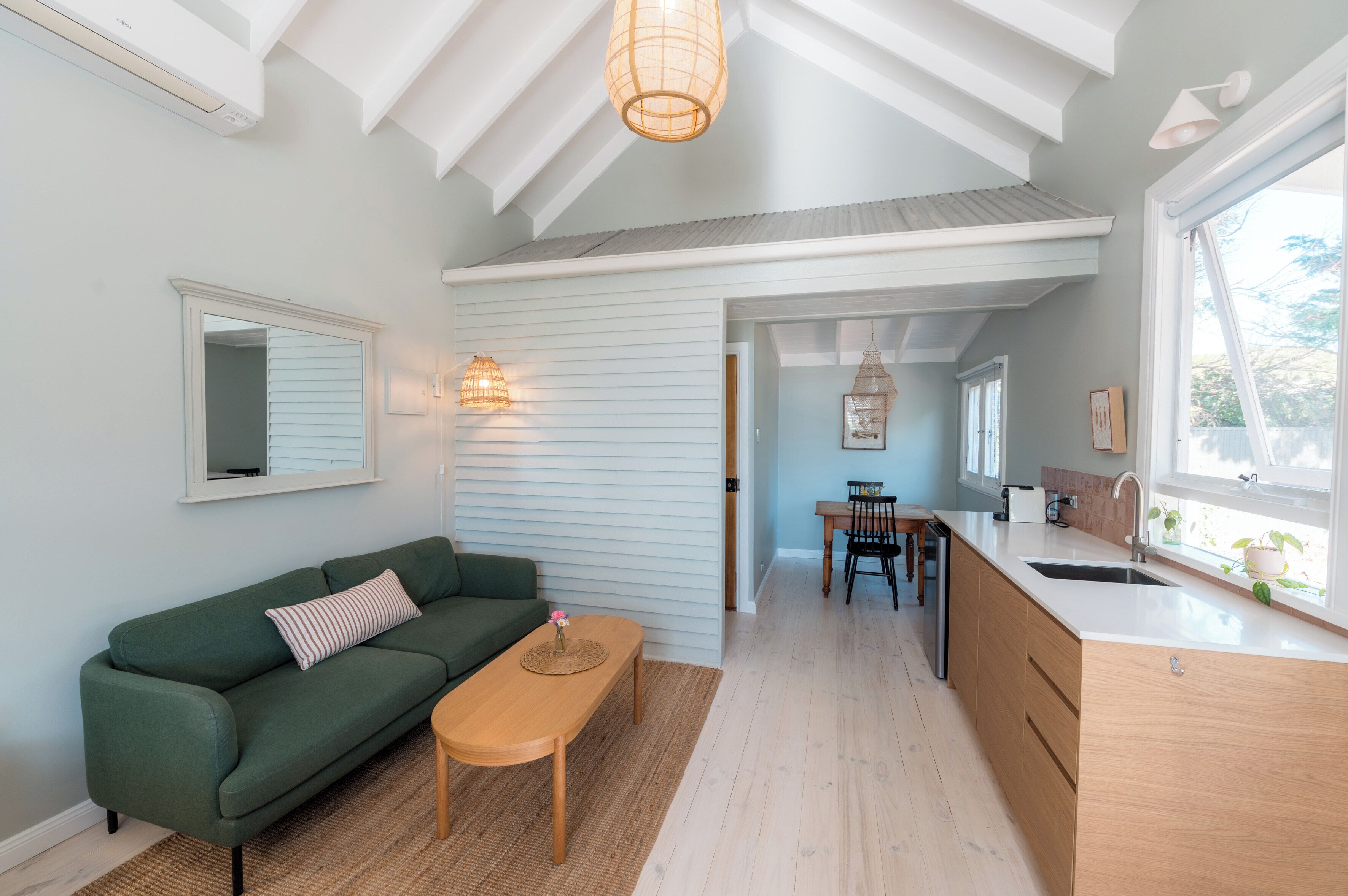 Una cocina moderna con encimera de madera y un fregadero, un comedor con mesa y sillas, y un sofá verde con un cojín estampado.