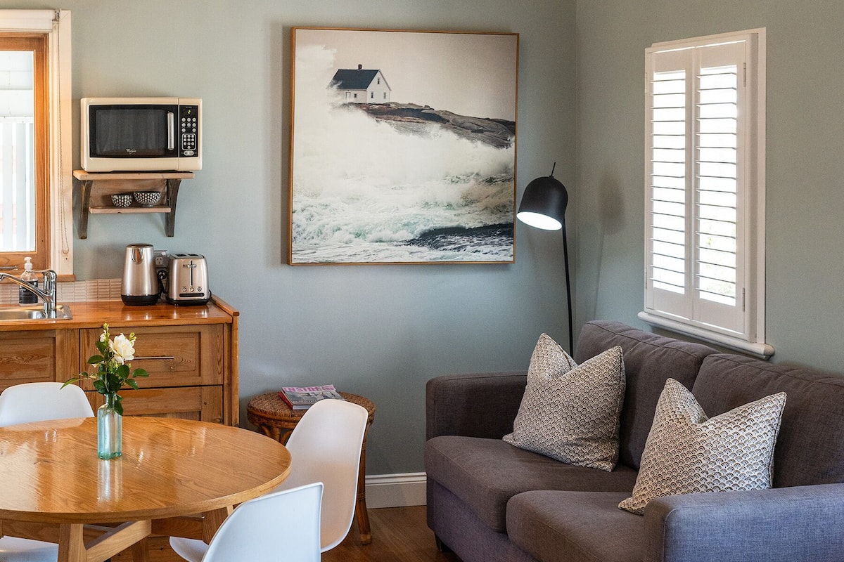 A cozy living room with a grey sofa, white chairs, a wooden dining table, and a painting of a ship on the wall.