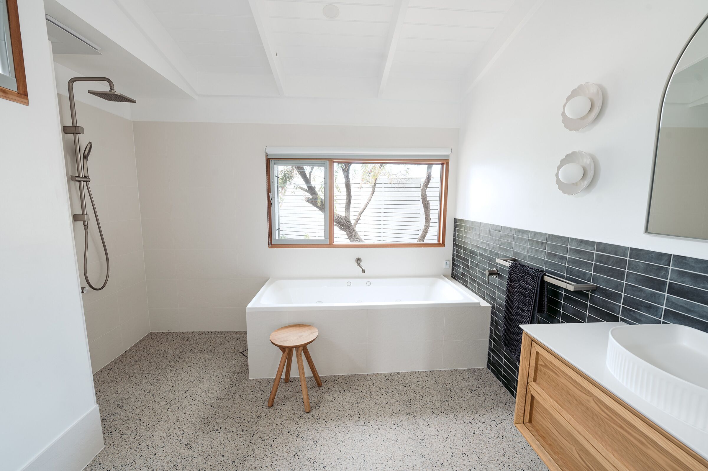A modern bathroom with a freestanding bathtub, a wooden stool, a large mirror, and a window with a view of trees.