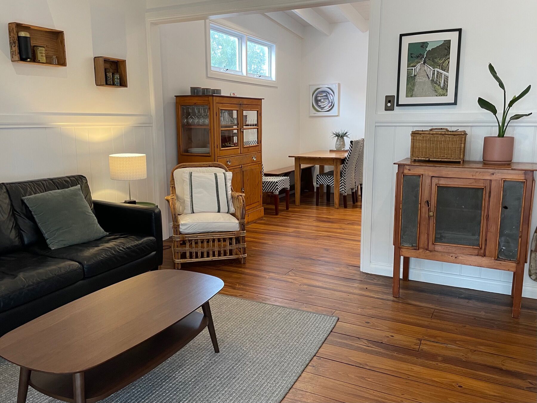 Una sala de estar acogedora con un sofá de cuero oscuro, una mesa de centro de madera y un sillón de mimbre. Se observa un aparador de madera con puertas de cristal y una zona de comedor con mesa y sillas.