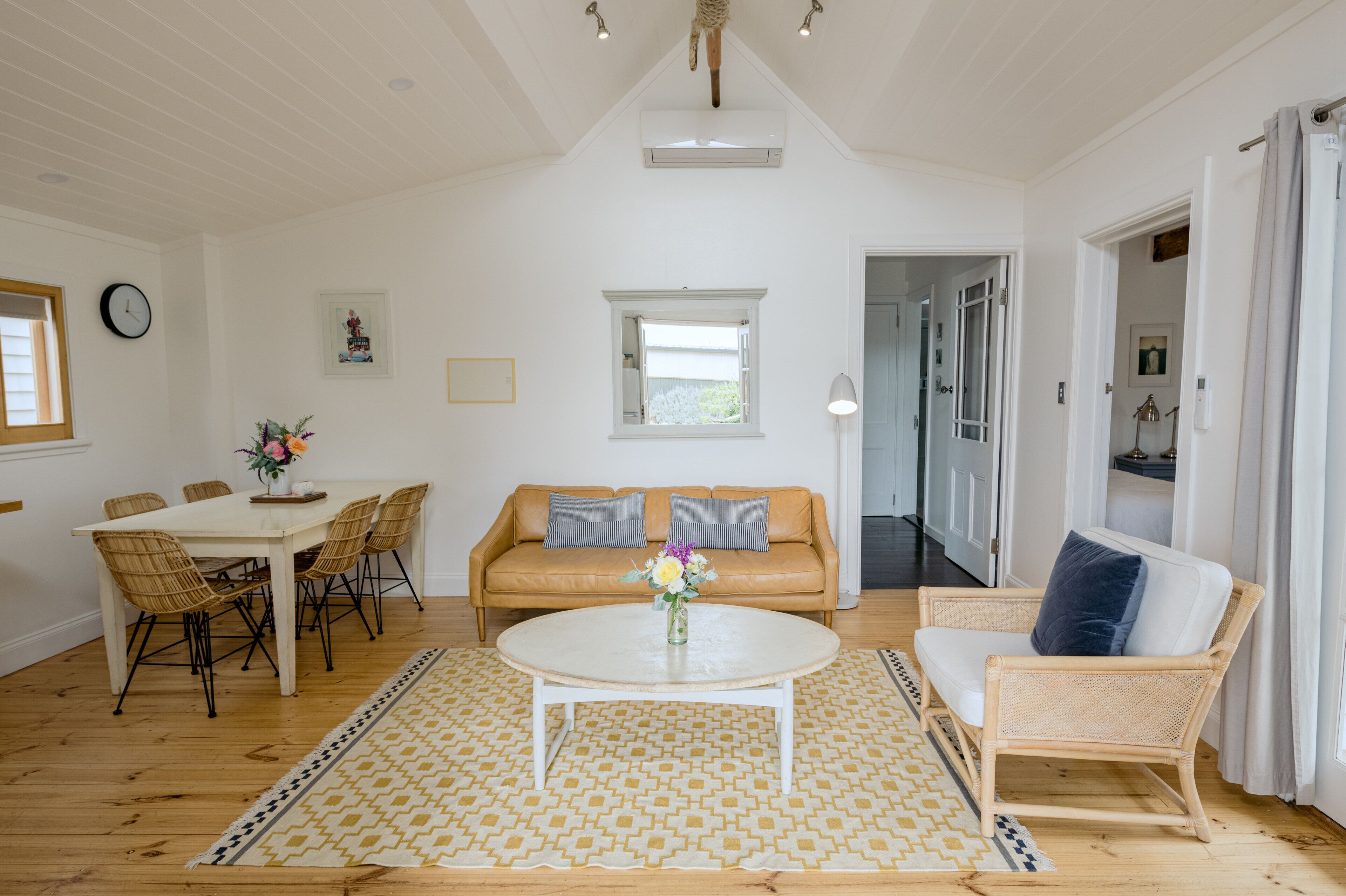 A spacious living room with a white ceiling, a dining area with wicker chairs, a sofa with cushions, and a coffee table with flowers.