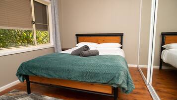 A single bed with a wooden frame and a green blanket, a window with blinds, and a view of greenery outside.