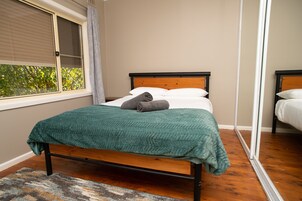 Una cama individual con estructura de madera y una manta verde, una ventana con persianas y vistas al exterior con vegetación.