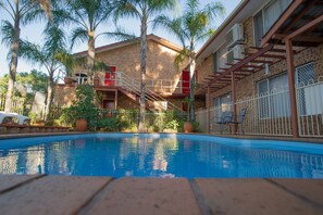 A swimming pool with a brick deck, surrounded by palm trees and a building with a red balcony. - Manera Heights Apartment Motel (Dubbo)