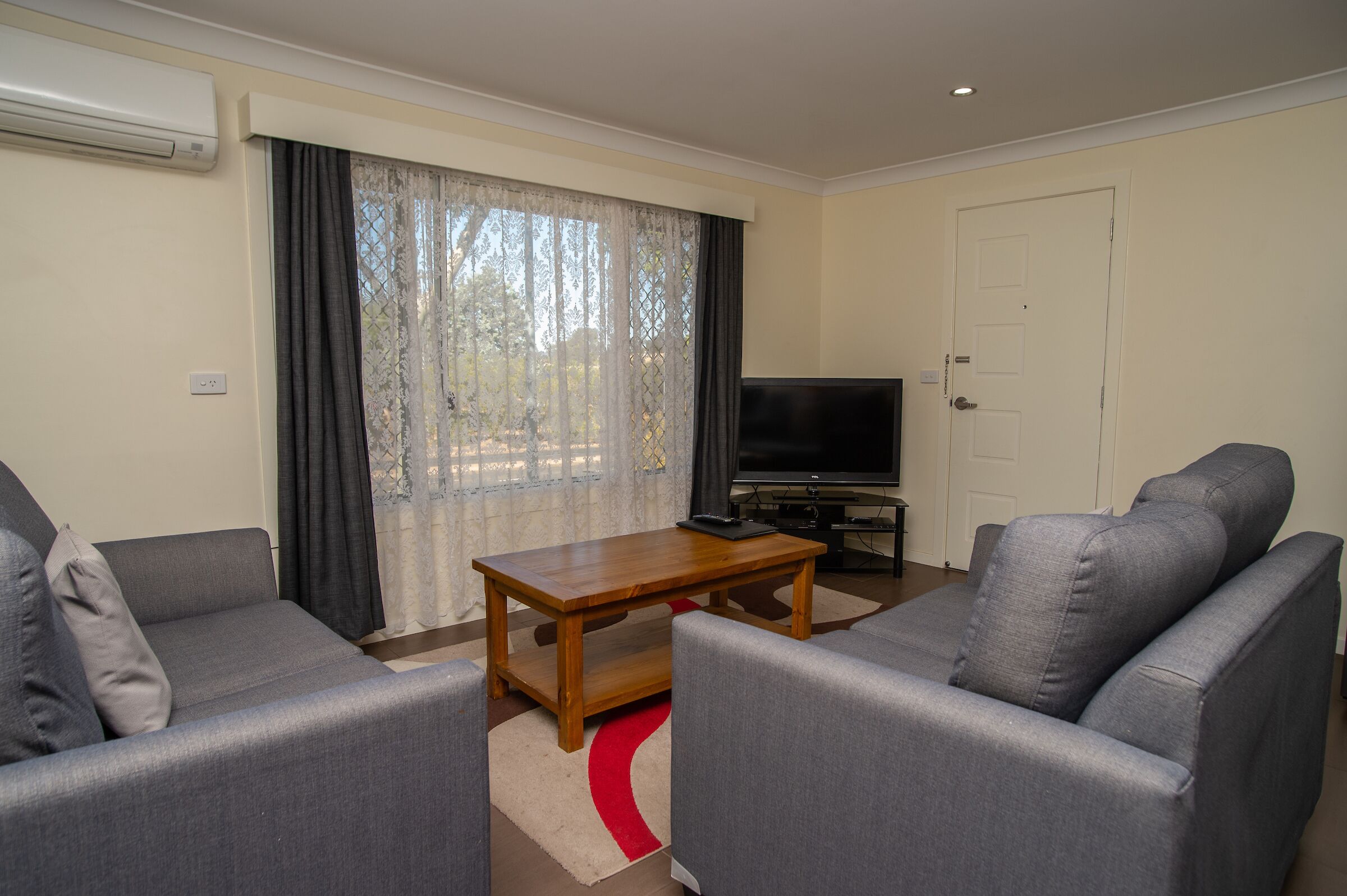 A living room with two grey sofas, a wooden coffee table, a flat-screen TV, and a large window with sheer curtains.