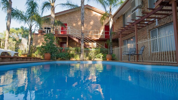 A swimming pool with a clear blue water surface, surrounded by a brick building with a balcony and a staircase leading to an upper level. - Manera Heights Apartment Motel (Dubbo)