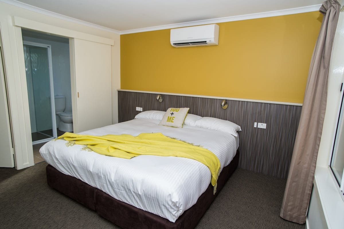 Habitación de hotel con cama, pared amarilla, unidad de aire acondicionado y baño con puerta de vidrio.
