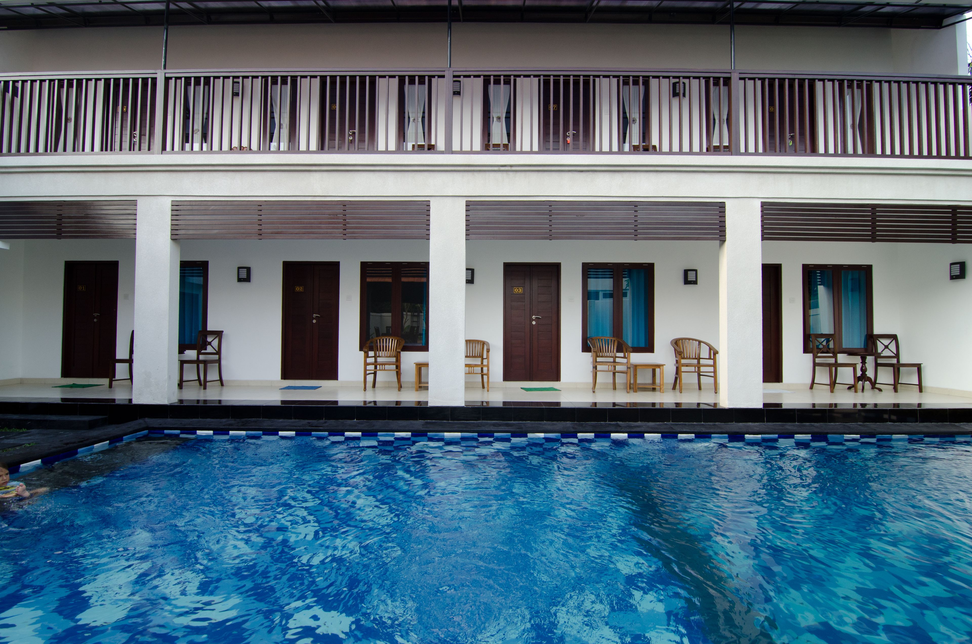 A swimming pool with a clear blue water surface, surrounded by a white building with multiple doors and windows, and a balcony with a railing.