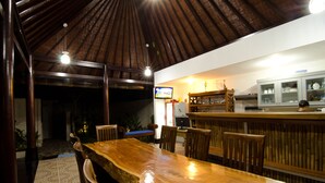 A restaurant with a wooden bar and seating area, a television, and a kitchen in the background. - Sanur Guest House (Denpasar)