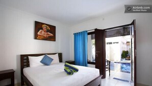 A hotel room with a bed, a nightstand, a painting on the wall, and an open door leading to a pool area. - Sanur Guest House (Denpasar)
