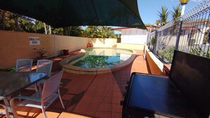 A swimming pool with a 'Shallow End' sign, surrounded by a tiled patio, metal chairs, and a covered area with a large umbrella. - Aspley Sunset Motel (Aspley)
