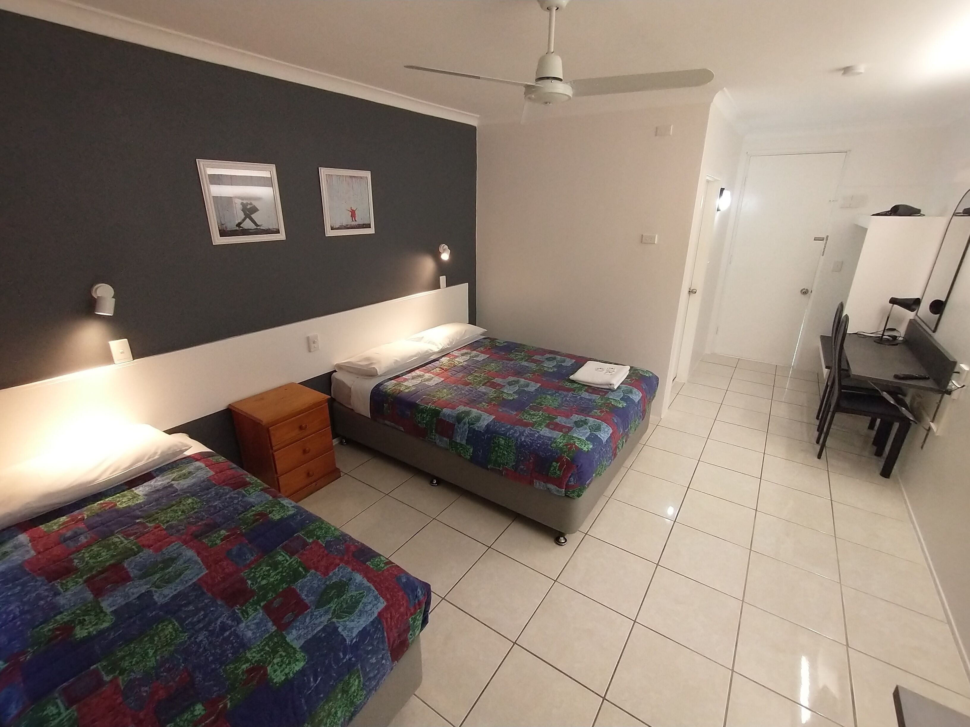 A bedroom with two beds, a ceiling fan, a desk with a chair, and a door leading to another room.