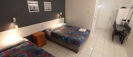 A bedroom with two beds, a ceiling fan, a desk with a chair, and a door leading to another room.