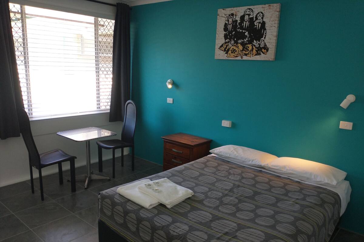 A bedroom with a bed, a desk, a chair, and a window with blinds.