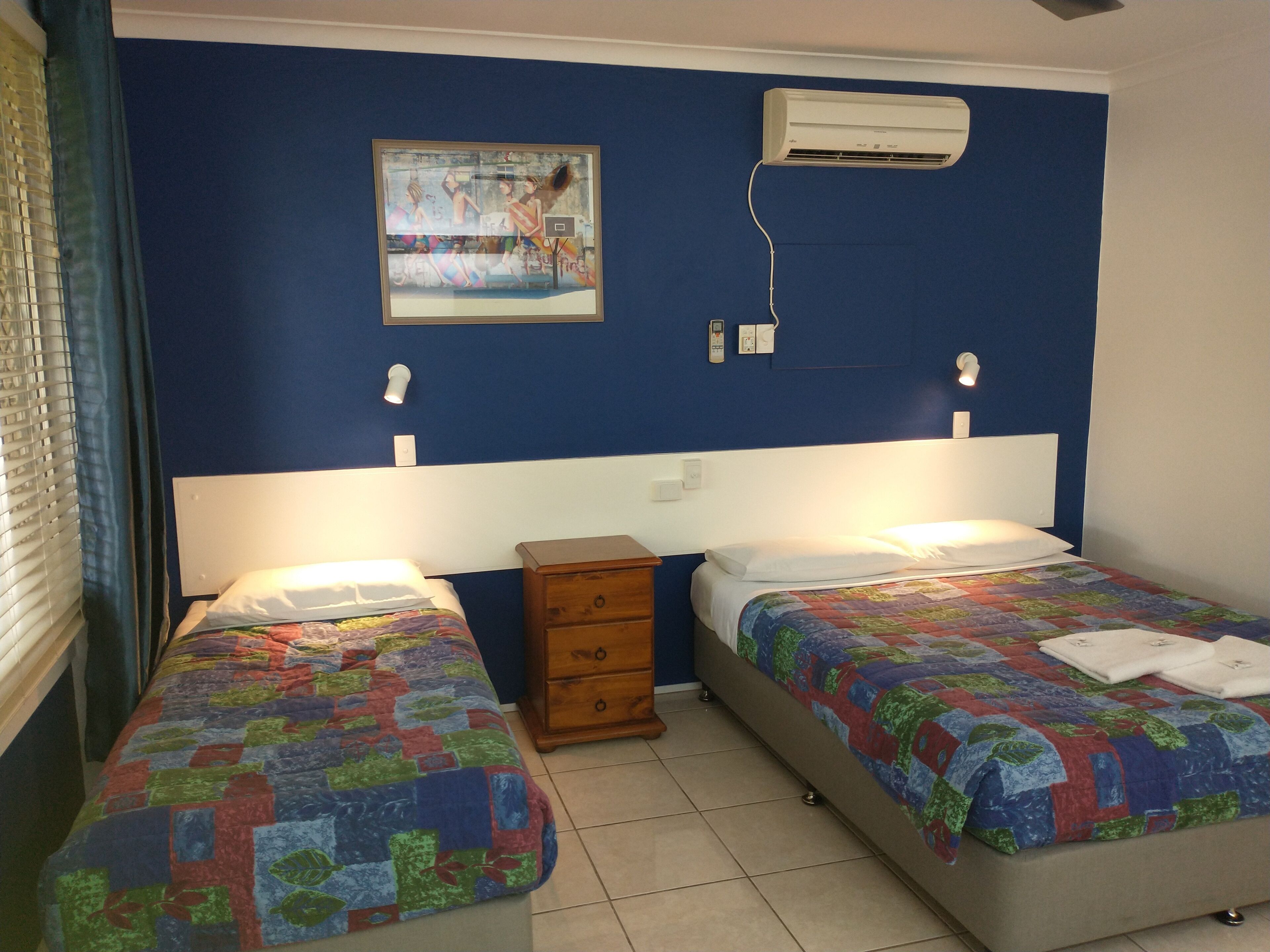 A room with two beds, a small wooden nightstand, a wall-mounted air conditioner, and a painting above the beds.