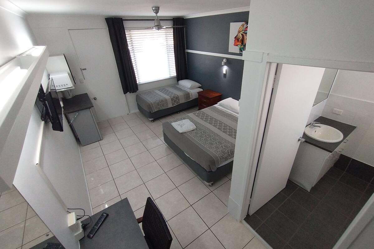 A compact hotel room with a bed, a desk, a chair, a small bedside table, a window with curtains, a ceiling fan, and a bathroom with a sink and mirror.