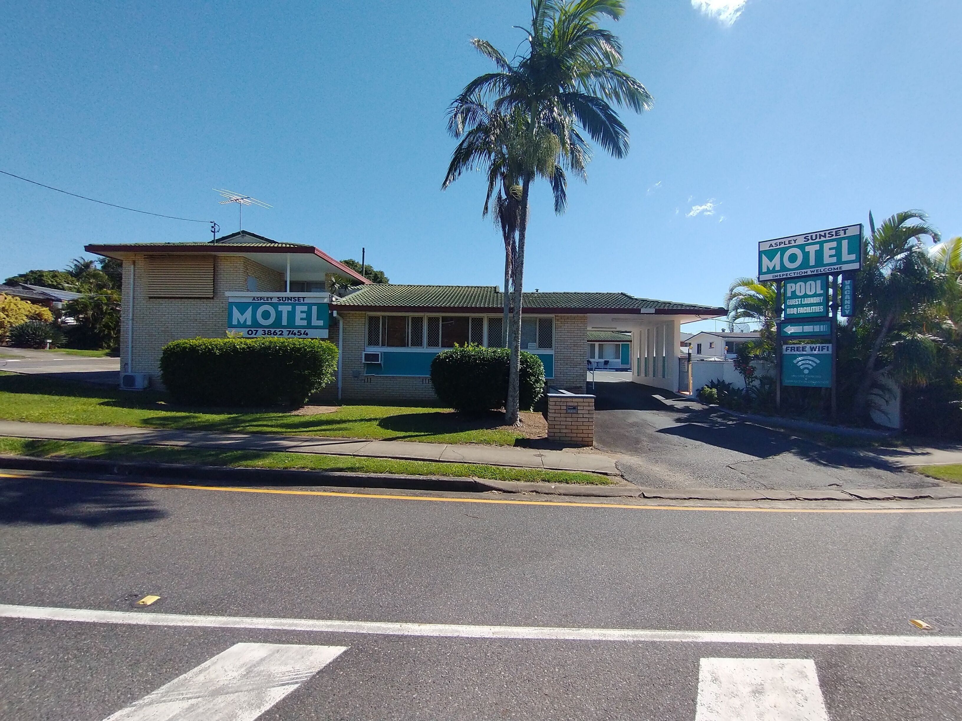 Un motel con una edificación de un solo piso, zona de estacionamiento techada y una palmera al frente.