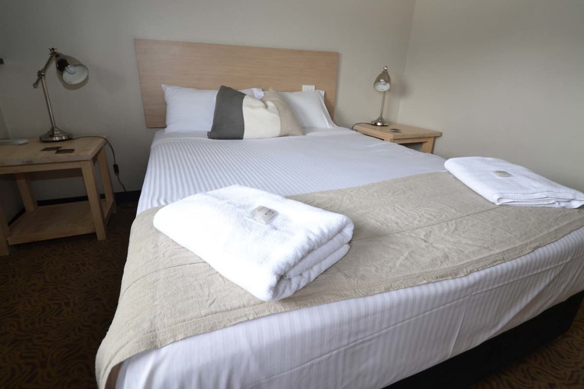 A neatly made bed with white linens and a folded towel on the side. There are two bedside tables with lamps.