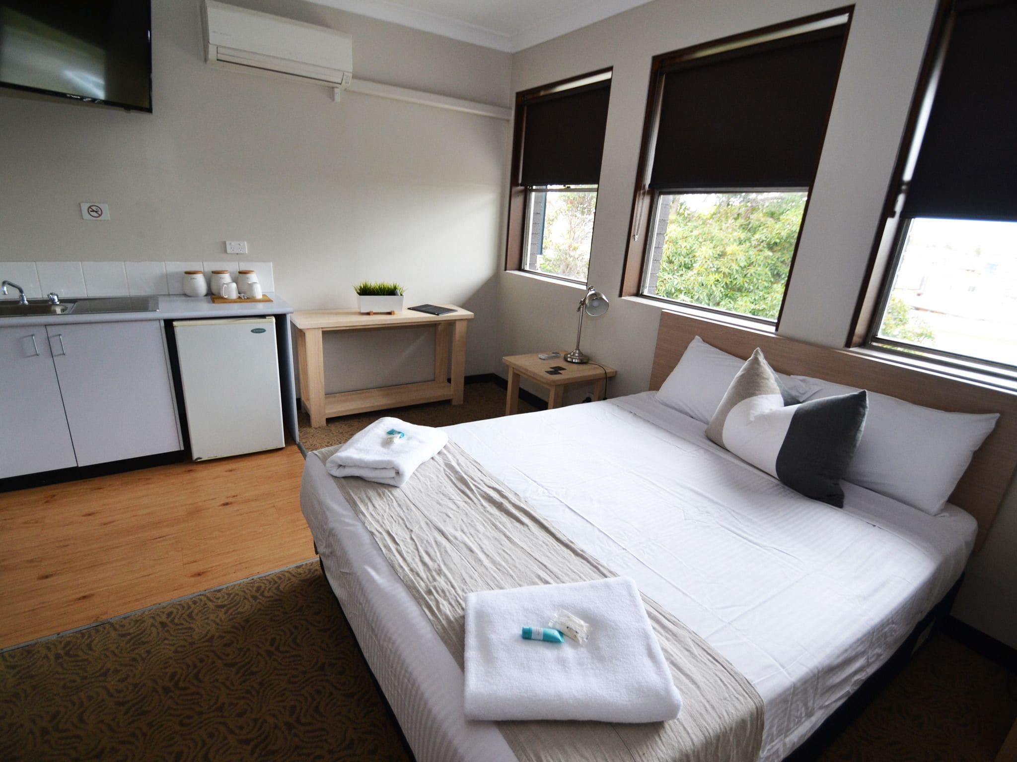 Habitación de hotel con cama, escritorio, silla, pequeña mesa, lámpara, televisor y zona de cocina.