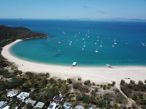 An aerial view of a coastal area with a sandy beach, several boats, and a few buildings with solar panels.