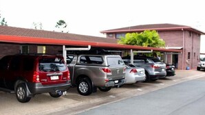 A parking area with several vehicles, including a red SUV and a silver SUV, parked in front of a building with a red roof and brick walls. - Motel Northview Mackay (Mount Pleasant)
