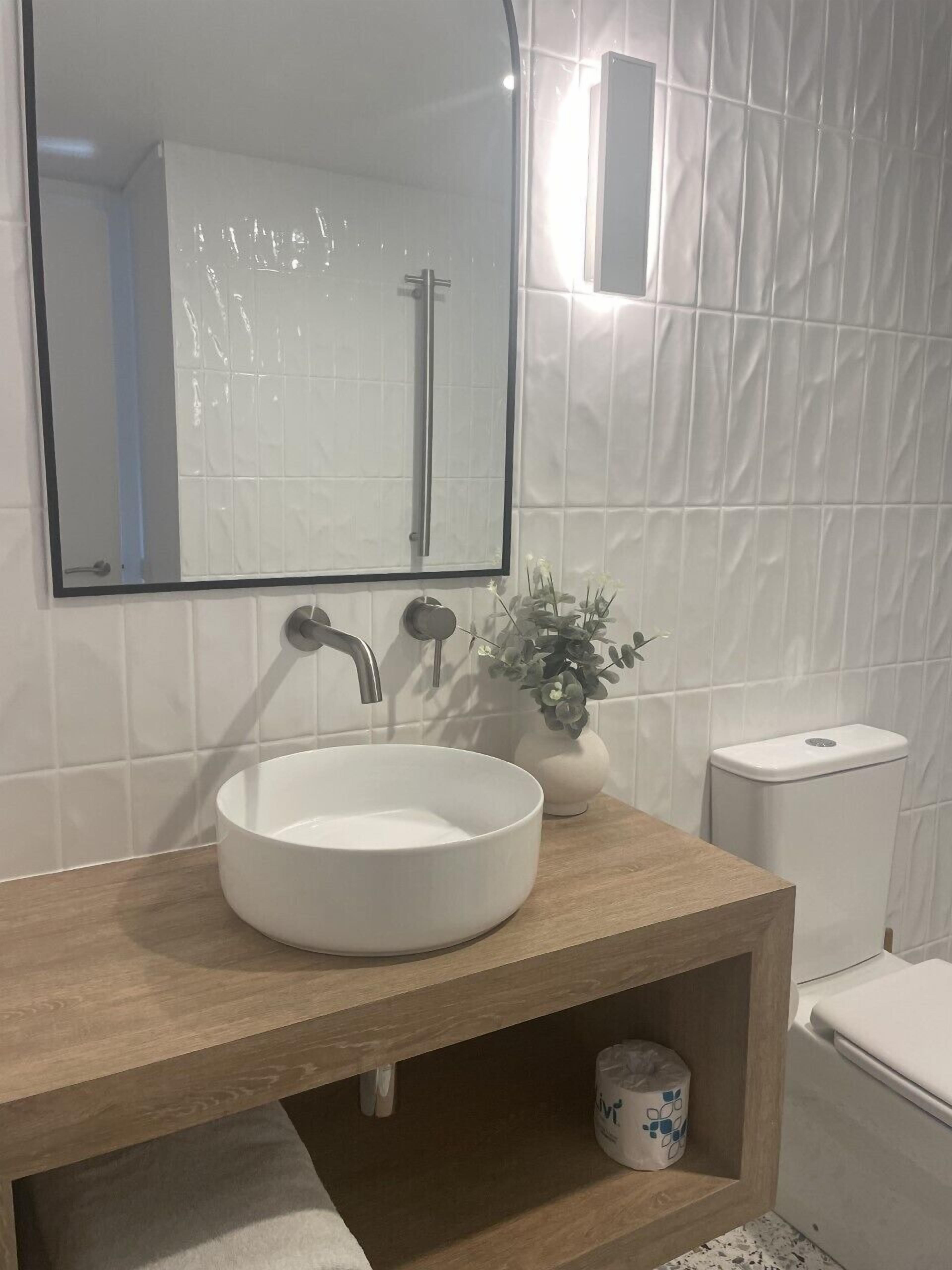 Une salle de bain avec un lavabo blanc, un meuble-lavabo en bois, un miroir, des toilettes et un luminaire fixé au mur.