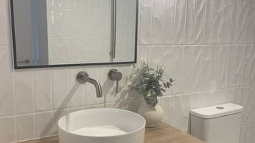Une salle de bain avec un lavabo blanc, un meuble-lavabo en bois, un miroir, des toilettes et un luminaire fixé au mur.