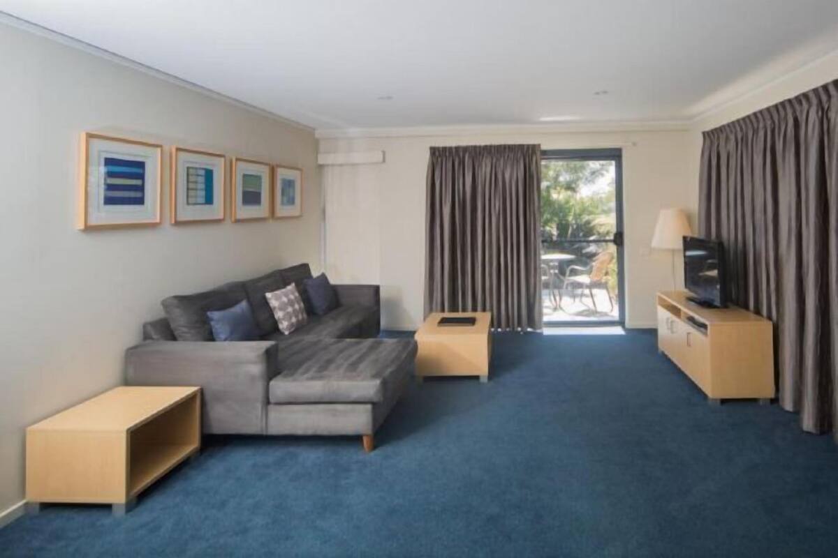 A modern hotel room with a grey sofa, a TV on a wooden stand, a small wooden side table, and a large window with curtains.