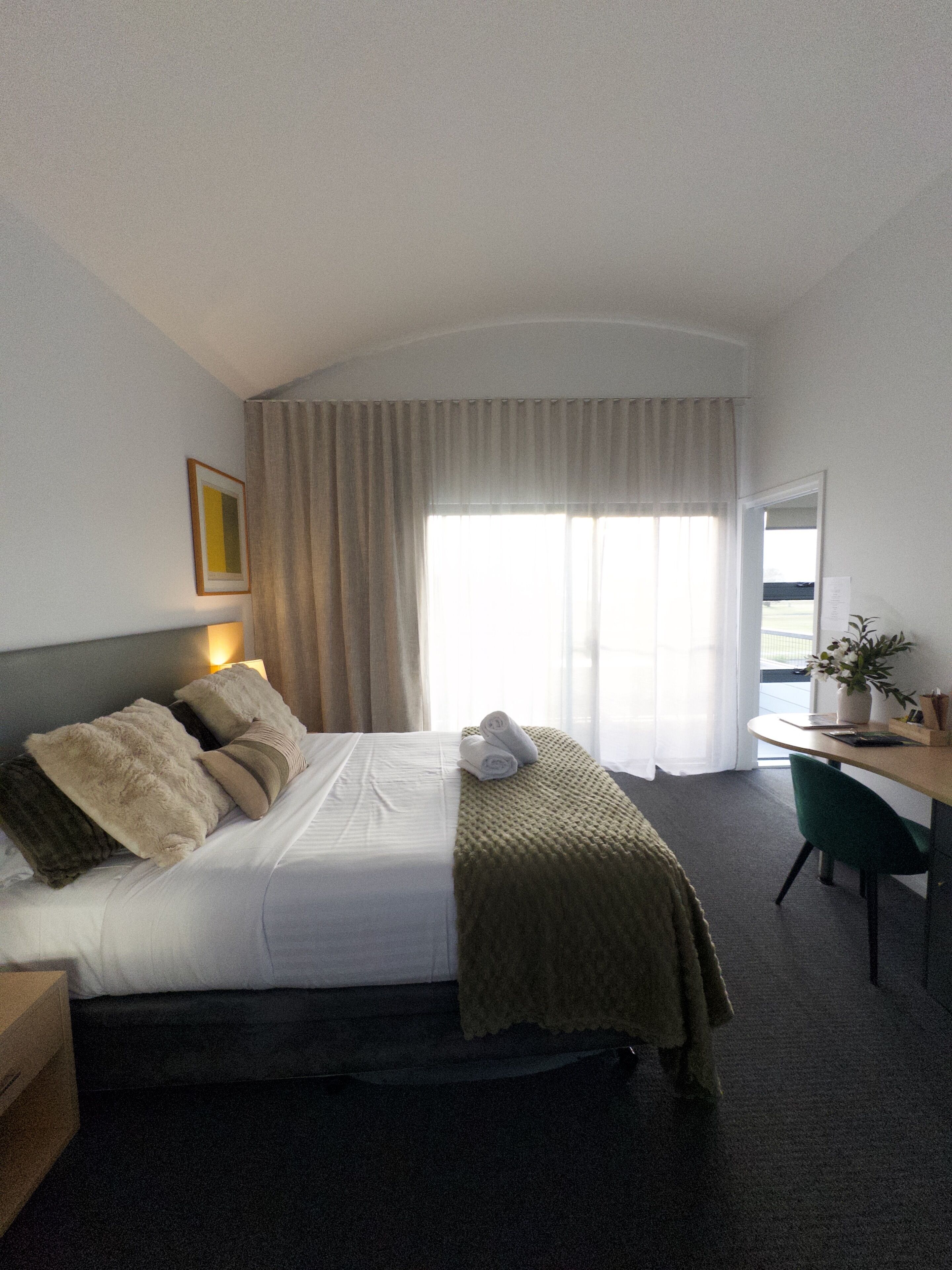 A hotel room with a bed, a desk, a chair, a window with curtains, and a plant.