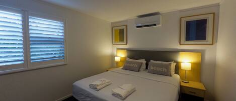 Une chambre d’hôtel avec un lit, deux lampes de chevet, une fenêtre avec des stores, un appareil de climatisation et un tableau encadré au mur.
