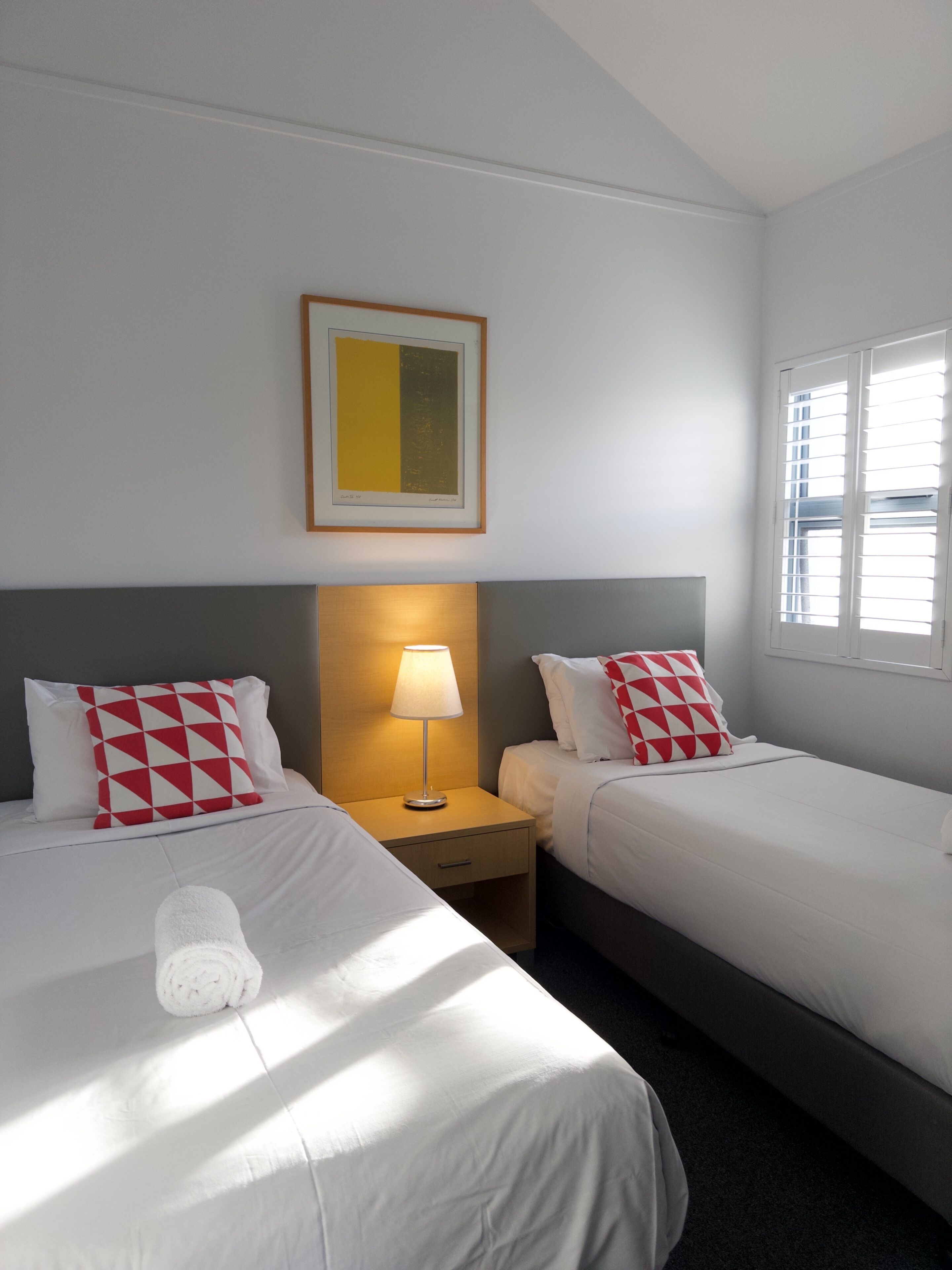 A hotel room with two beds, a nightstand with a lamp, a framed picture on the wall, and a window with blinds.