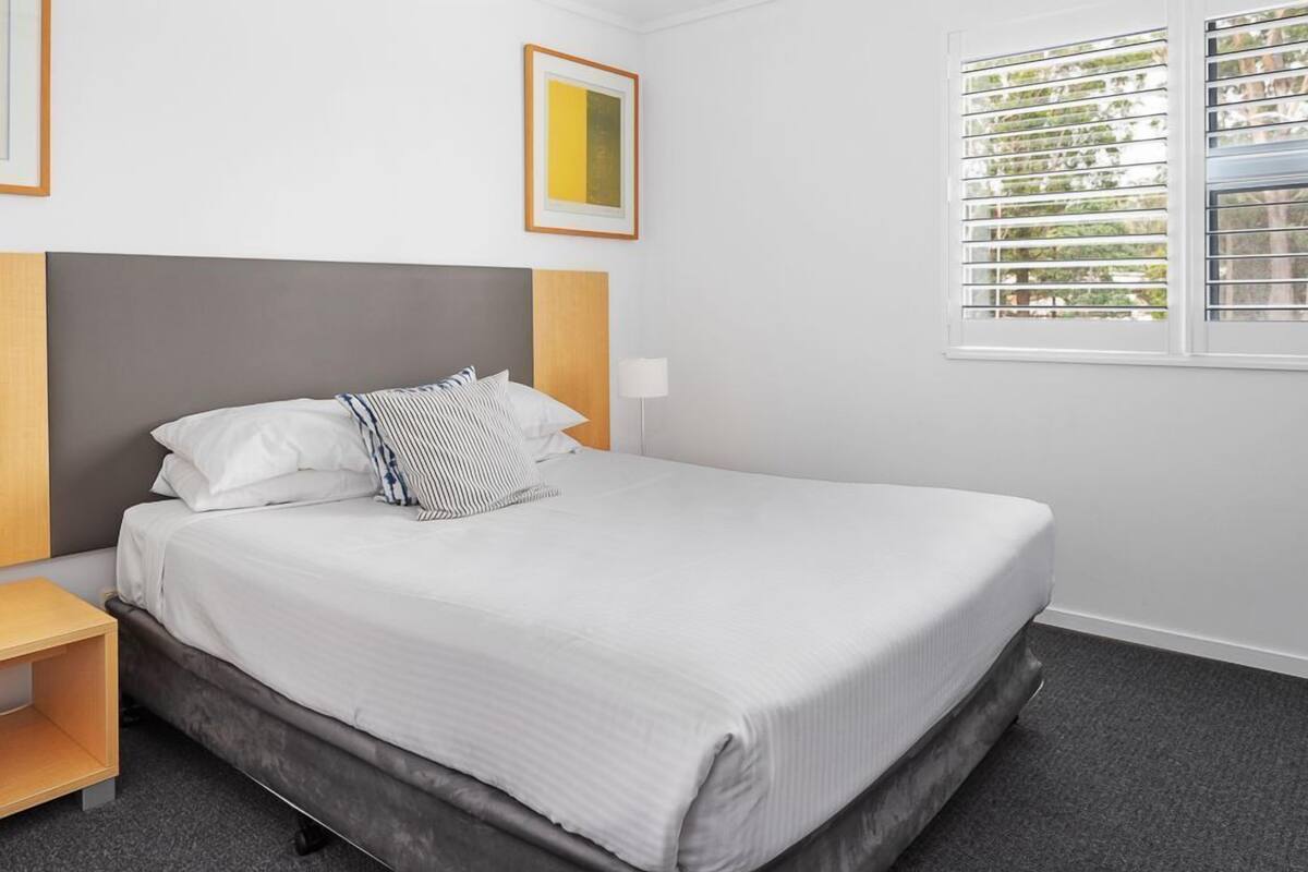 A neatly made bed with white bedding and striped pillows, a wooden nightstand, a lamp, and a window with blinds.