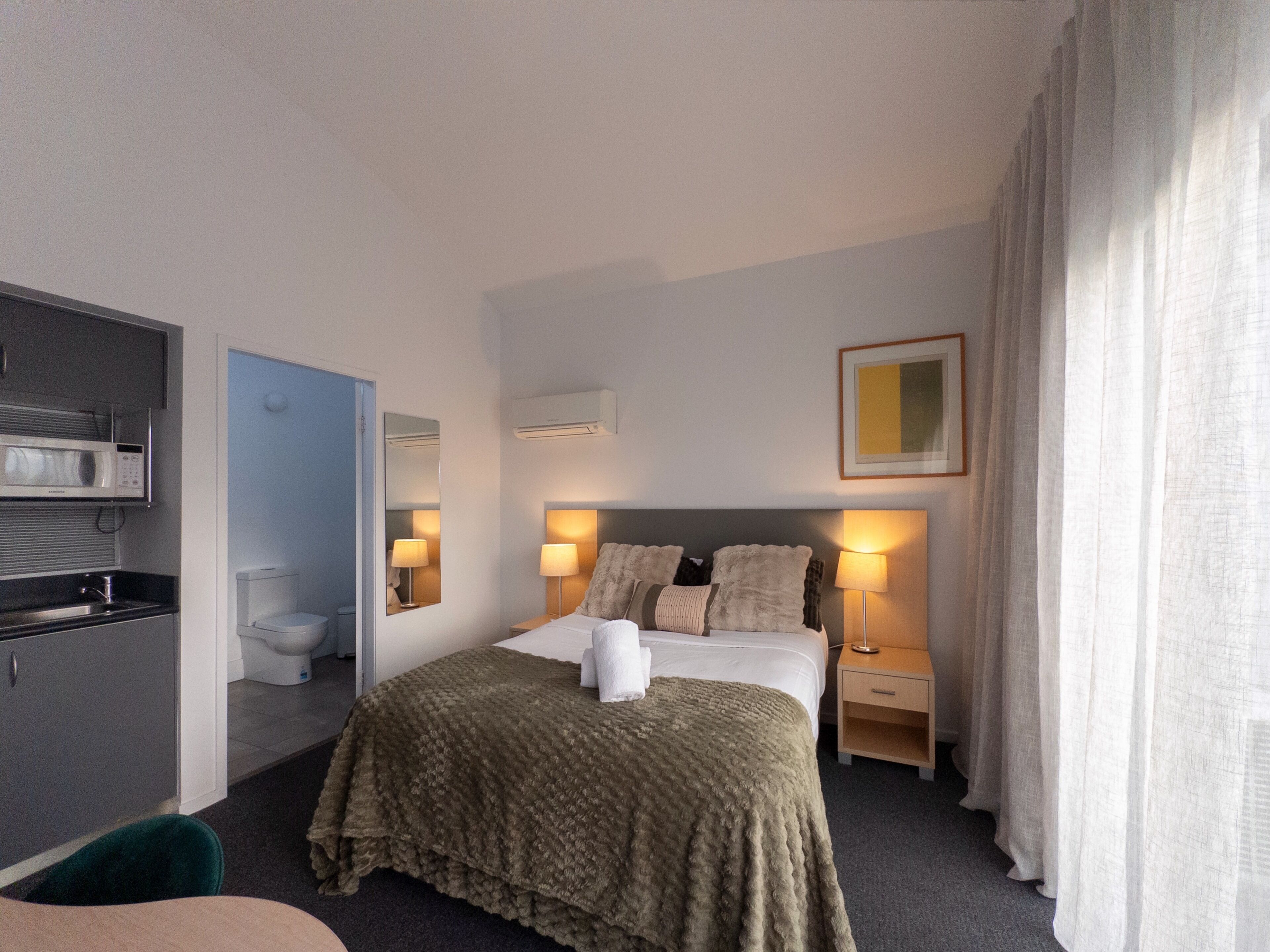 Ett hotellrum med en säng, sängbord, en spegel, en luftkonditioneringsanläggning, en mikrovågsugn och en toalett.