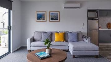 A modern living room with a grey sectional sofa, a coffee table with a book, and a potted plant. There are two framed pictures on the wall and a kitchen area with a microwave and refrigerator.