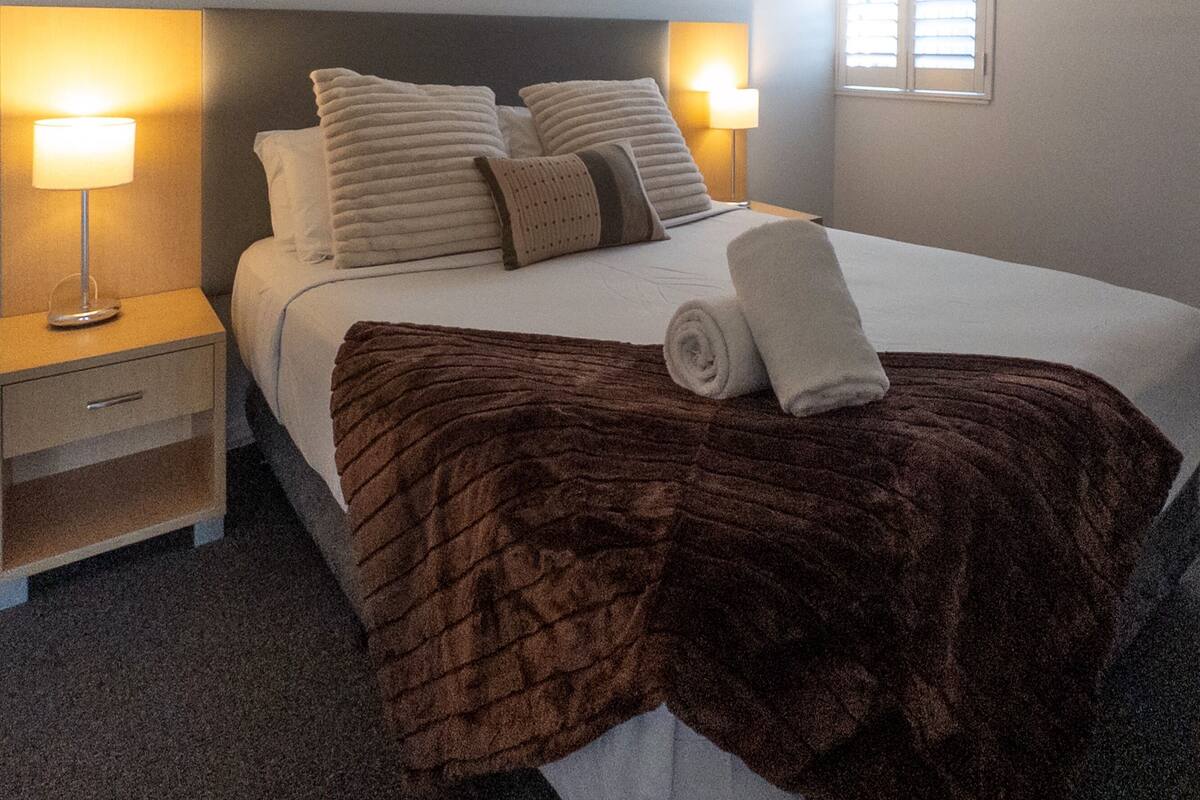 A hotel room with a bed, two bedside lamps, a framed picture on the wall, and a window with blinds.