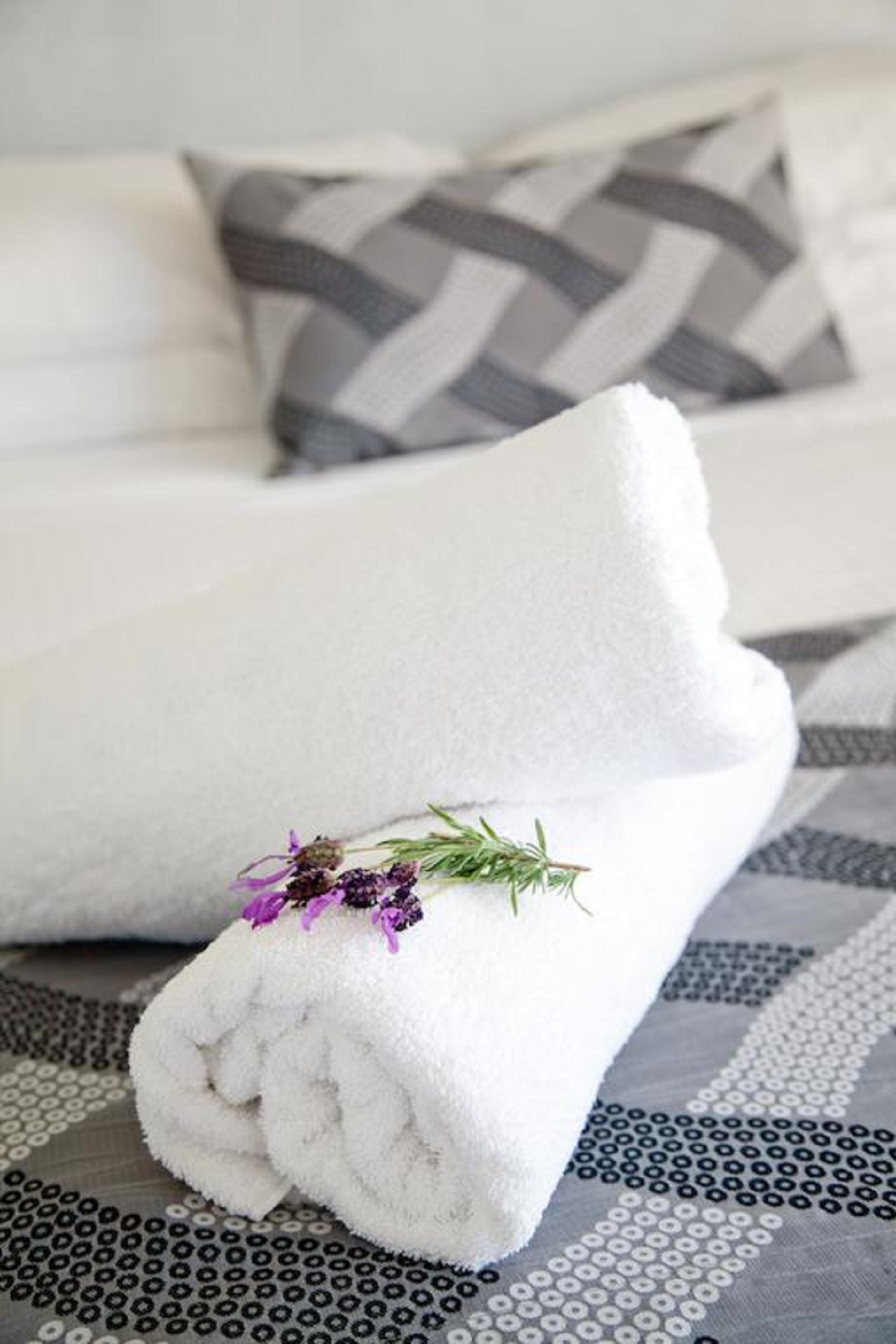 A neatly folded white towel with a sprig of rosemary and purple flowers on a patterned bedspread.