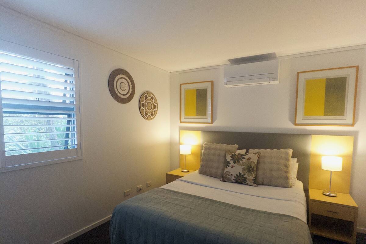 A bedroom with a bed, two bedside tables with lamps, a window with blinds, and wall decorations.