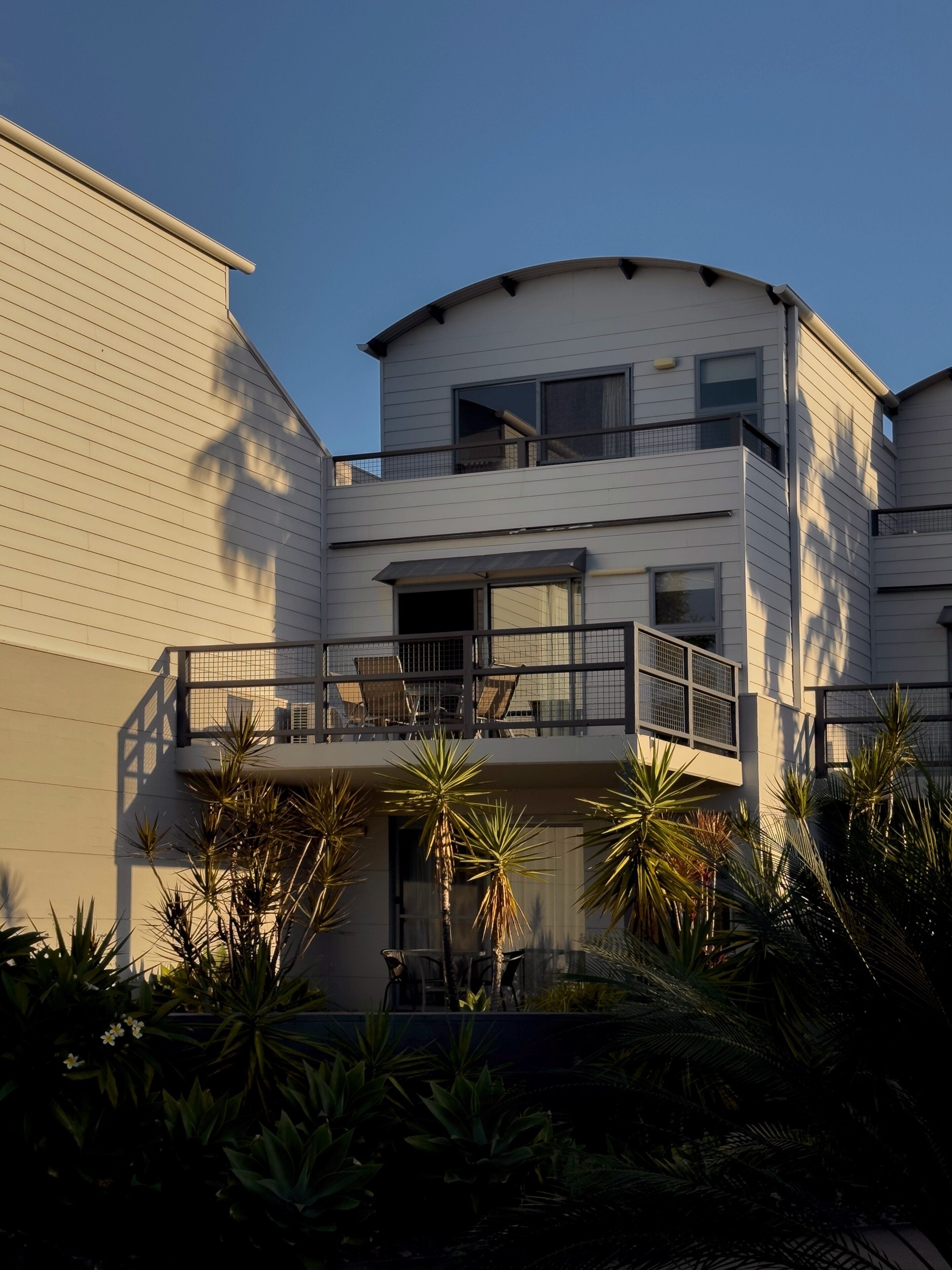 A multi-story residential building with balconies, large windows, and surrounding tropical plants.