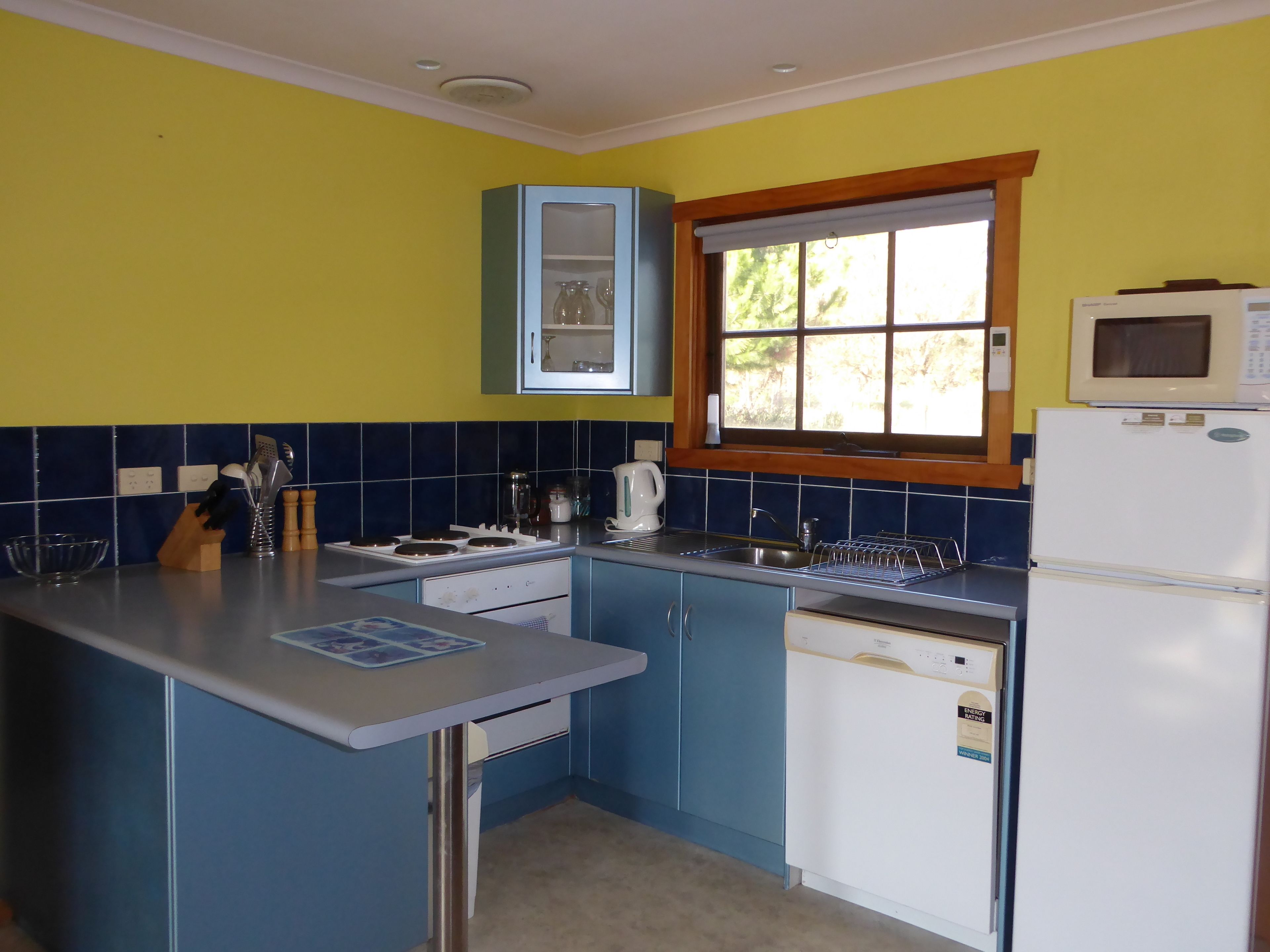 Une cuisine de taille réduite avec des meubles bleus, un réfrigérateur blanc et une fenêtre donnant sur un paysage verdoyant.