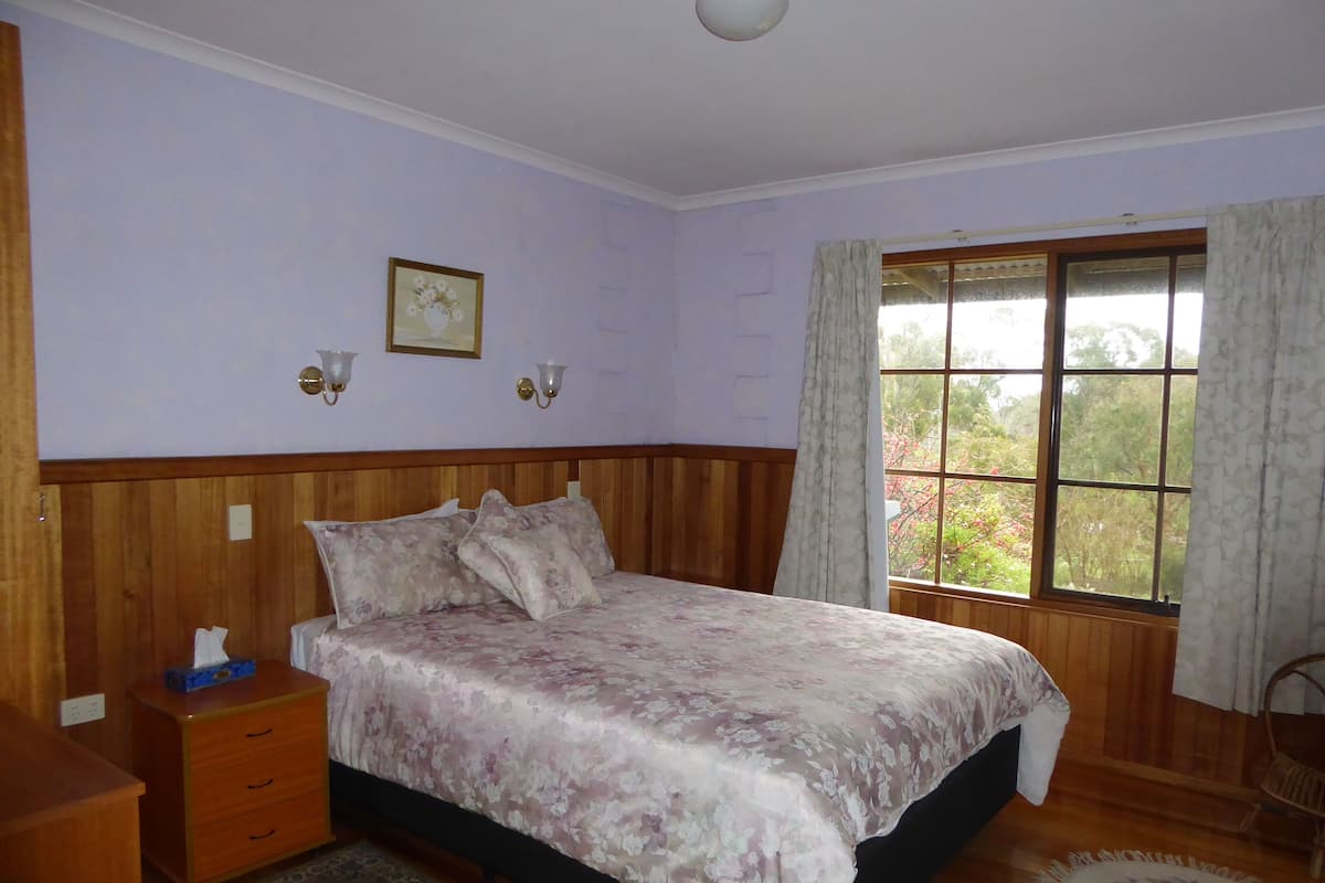 Ein Schlafzimmer mit einem hölzernen Kopfende, einer blumigen Bettdecke, einem Nachttisch mit Taschentuchbox, einem Beistelltisch, einem Fenster mit Vorhängen und Blick auf Bäume.