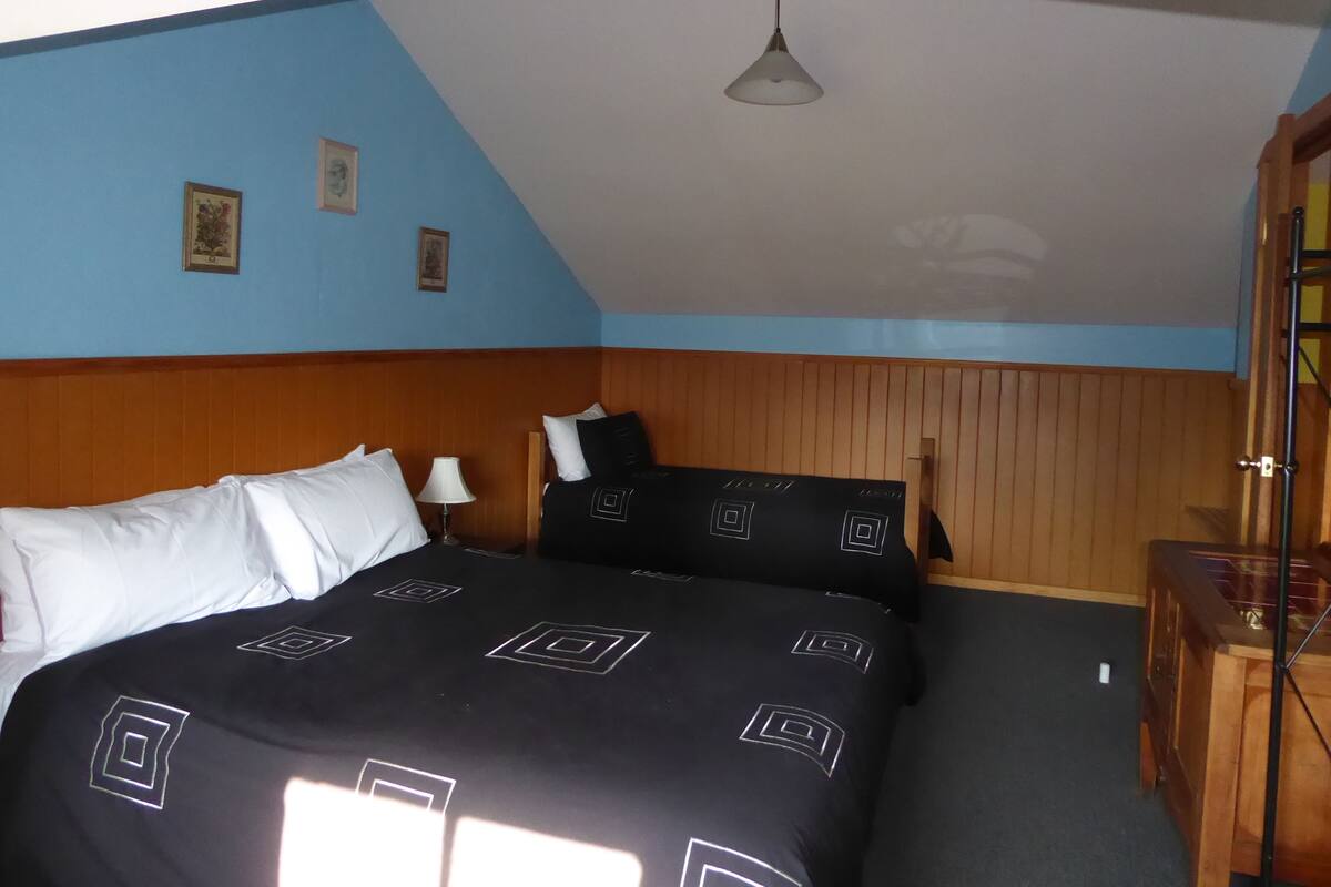 Ein Schlafzimmer mit schrägem Dach, einem Bett mit schwarz-weiß gestreifter Bettwäsche, einem Holz-Kopfende, einem Nachttisch mit Lampe und einem Holzschrank.