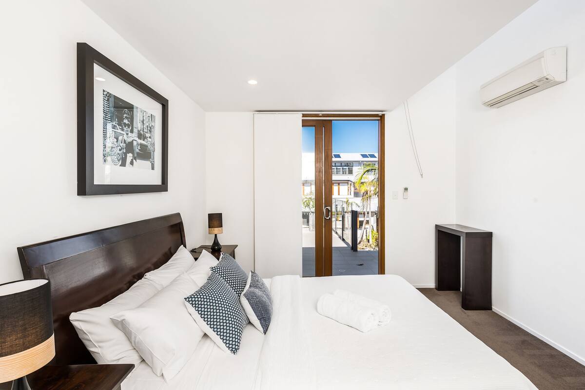 Un hotel moderno con una cama grande, un cuadro enmarcado en la pared, una mesita de noche con lámpara y vistas al exterior a través de una puerta corrediza.
