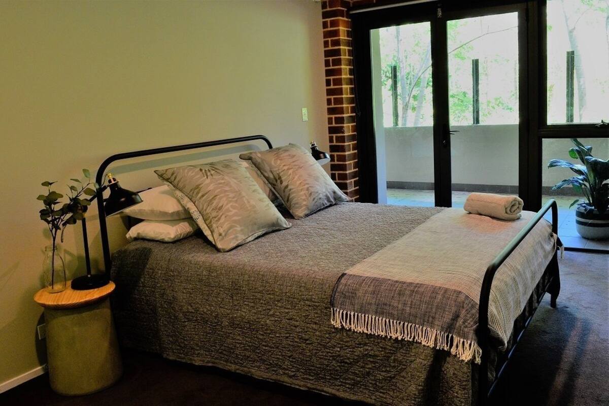 Una cama bien hecha con colcha gris, dos almohadas y una manta. Hay una mesita de noche con una lámpara y una planta. La habitación tiene un ventanal con vistas a árboles.