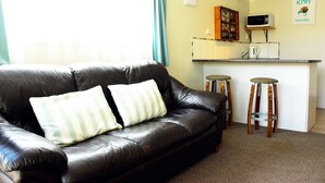 A living room with a black leather sofa, two wooden stools, a kitchen area with a microwave, and a framed picture on the wall. - Te Awa Motel (Whanganui)