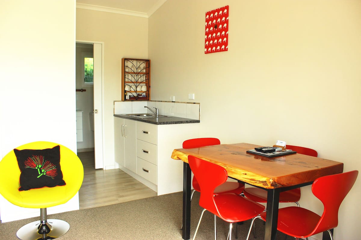 A dining area with a wooden table and red chairs, a yellow chair with a black cushion, and a kitchen in the background.