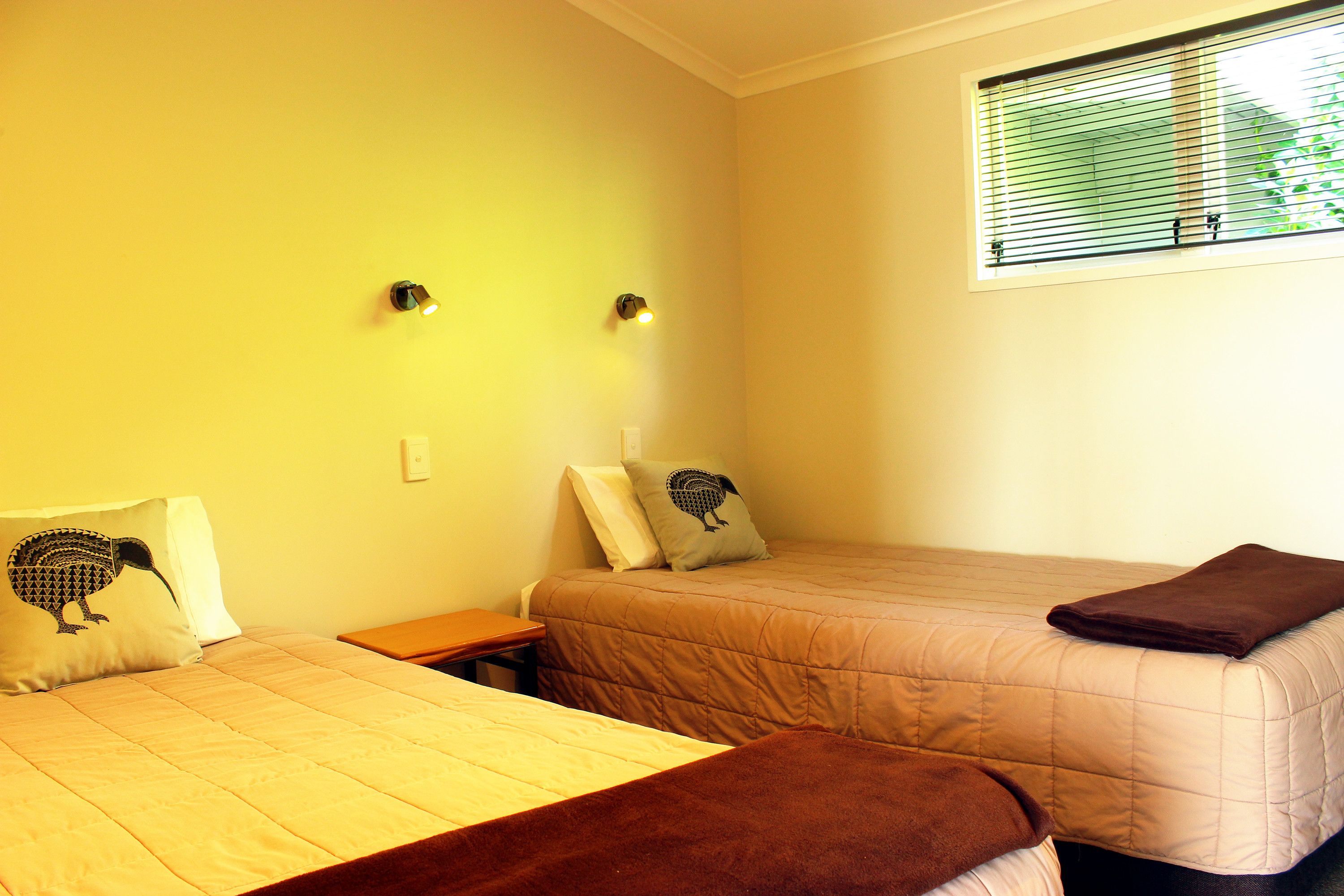 A bedroom with two beds, each with a pillow and a blanket. There is a window with blinds, a nightstand, and a wall-mounted light fixture.