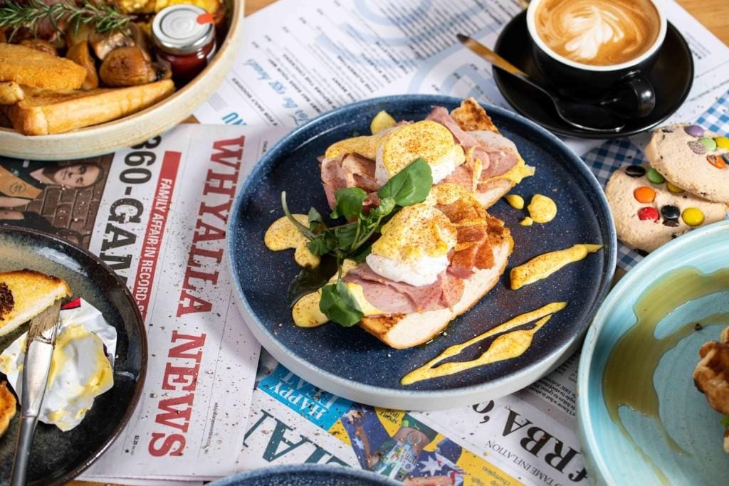 A breakfast setting with a plate of eggs Benedict, a cup of coffee, and a newspaper spread out on the table.