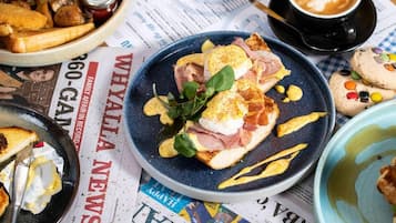 Uma mesa posta para o café da manhã, com ovos Benedict, uma xícara de café e um jornal aberto.